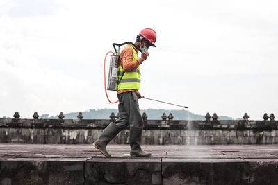 Side view of worker spraying chemical on footpath against sky