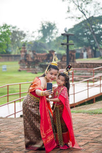 An asian woman in a red thai traditional dress takes a picture with her daughter on her phone