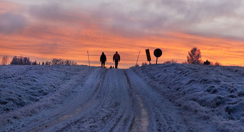 People walking on snow covered landscape against sky during sunset