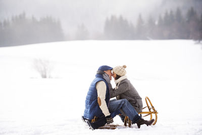 Happy senior couple sitting face to face on sledge in winter landscape