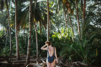 Back view of peaceful unrecognizable young female in swimwear standing with hands on head and eyes closed against tall green palms during summer holidays on seacoast of costa rica