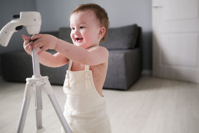 European child of 1 year in white jumpsuit with tripod for photo of house in bright real living room