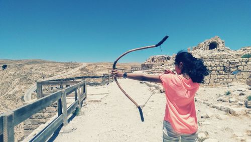 Rear view of woman aiming with bow and arrow by old ruins against clear sky