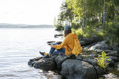Young woman in yellow hoodie with backpack sitting on rocks looking at view of lake enjoying nature 