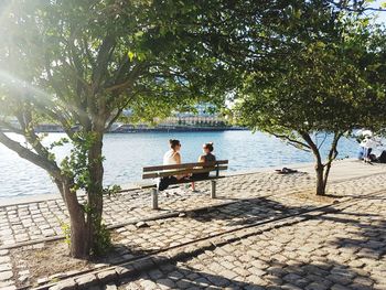 People sitting on riverbank by river against sky