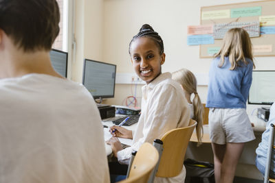 Side view portrait of smiling girl sitting on chair with friends in computer class at school