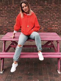 Full length portrait of woman sitting on picnic table against brick wall