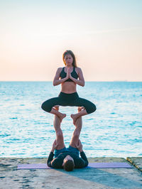 Couple practicing yoga on pier by sea against sky during sunset