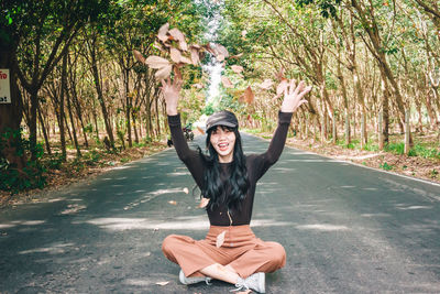 Portrait of smiling young woman throwing leaves while sitting on road amidst trees in forest