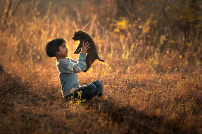 Side view of cute girl carrying puppy while sitting on field during sunset