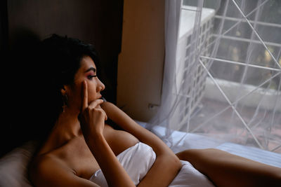 High angle view of thoughtful naked young woman with blanket sitting on bed at home