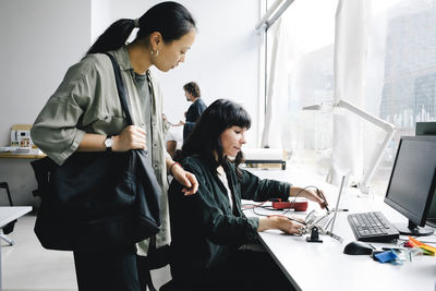 Female customer with tote bag standing near technician examining laptop at repair shop