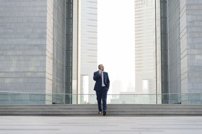 Full length of businessman talking on mobile phone while walking against office buildings