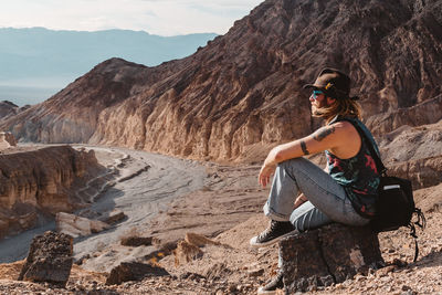 Male with hat and camera bag sitting on rocks in remote valley looking outward in death valley.