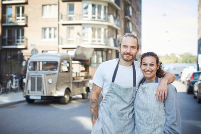 Portrait of confident male and female owners standing arms around with food truck in background