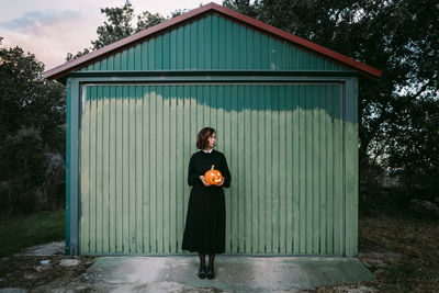 Female in black dress and with halloween pumpkin lantern standing against wooden wall of house and looking away