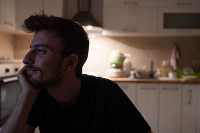 Young european guy pictured in his kitchen, pressing fist to jaw as if dreaming of something.