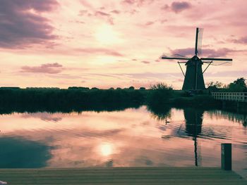 Traditional windmill by lake against cloudy sky during sunset