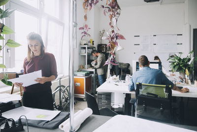 Creative businesswoman examining papers while colleagues working in background at office