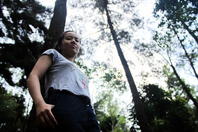 Low angle portrait of woman standing against tree in forest