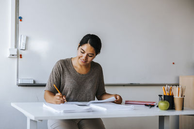 Smiling professor checking test papers while sitting near table in classroom at school