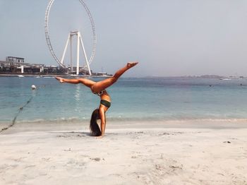 Full length of woman practicing handstand on shore at beach against sky