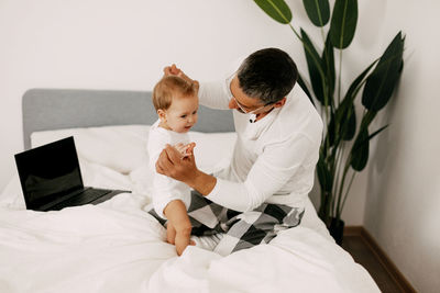 Dad sits on the bed, works on a laptop at home with a baby on his lap, smiles 