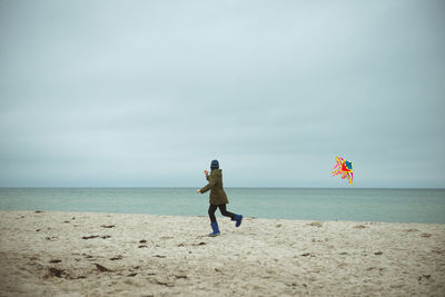 Rear view of boy holding kite running on beach against sky