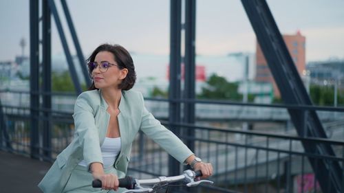 Young casual businesswoman enjoys riding bike in city on bridge with beautiful cityscape