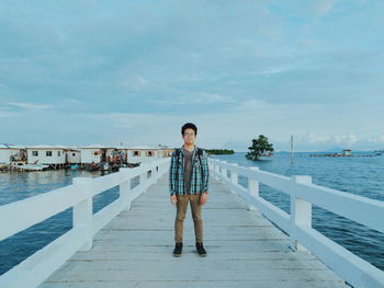 Full length of young man standing on footbridge over sea against sky