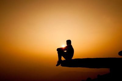 Silhouette of man sitting on cliff against sky during sunset