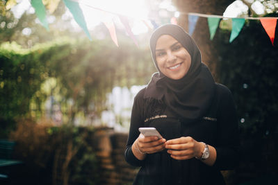 Portrait of smiling woman in hijab holding smart phone at backyard