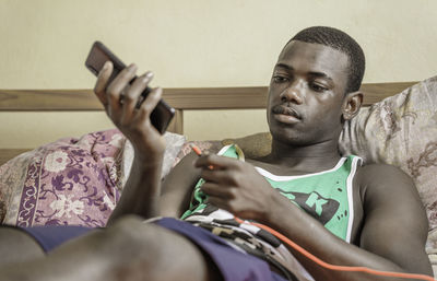 Focused african american male with short hair lying on bed and plugging usb cable into mobile phone