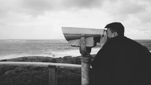 Side view of man looking through coin-operated binoculars at beach against sky