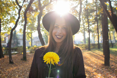 Portrait of smiling young woman holding flower while standing against trees