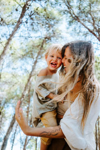 From below side view of positive mother with adorable little kid having fun and laughing together in woods during summer weekend