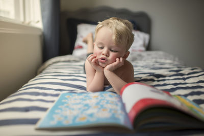 Boy with hands on chin reading picture book while lying on bed at home