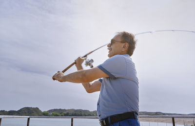 Senior man fishing with fishing rod by sea on sunny day