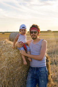 Father and son in t-shirts standing next to a haystack on a sloping field during sunset