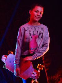 Portrait of girl with skateboard standing against sky at night