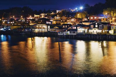 Illuminated buildings by river in town against sky at night