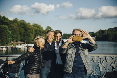 Smiling male and female friends taking selfie with smartphone by railing of bridge