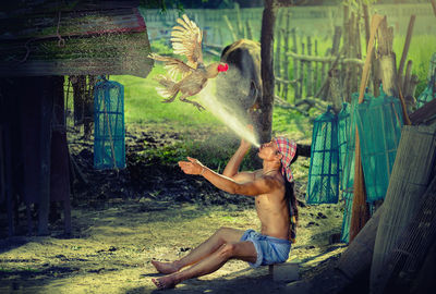 Full length of shirtless man spraying water on flying rooster
