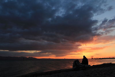 Silhouette sitting on beach by sea against sky during sunset