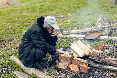 An elderly village man is sitting with a cigarette and lighting a fire on the street near the house