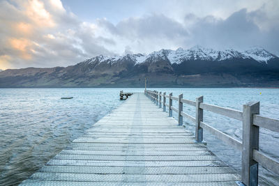 Wide scenic view of pier over calm lake against snowcapped mountain range