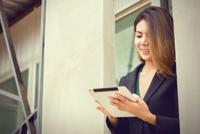 Portrait of young woman using phone while standing against wall