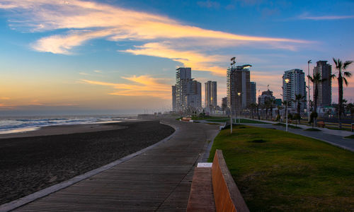 Footpath by sea and buildings against sky during sunset