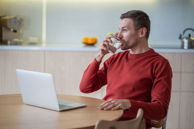 Freelancer working from home using laptop in cozy modern apartment, drinking water during break