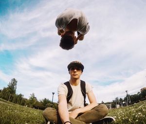 Man jumping over friend sitting on grass against sky